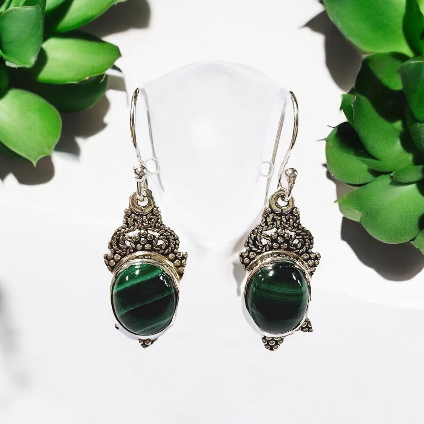 A pair of silver earrings featuring intricate Persian-inspired designs and malachite gemstones, displayed on a white stand with a green plant in the background.