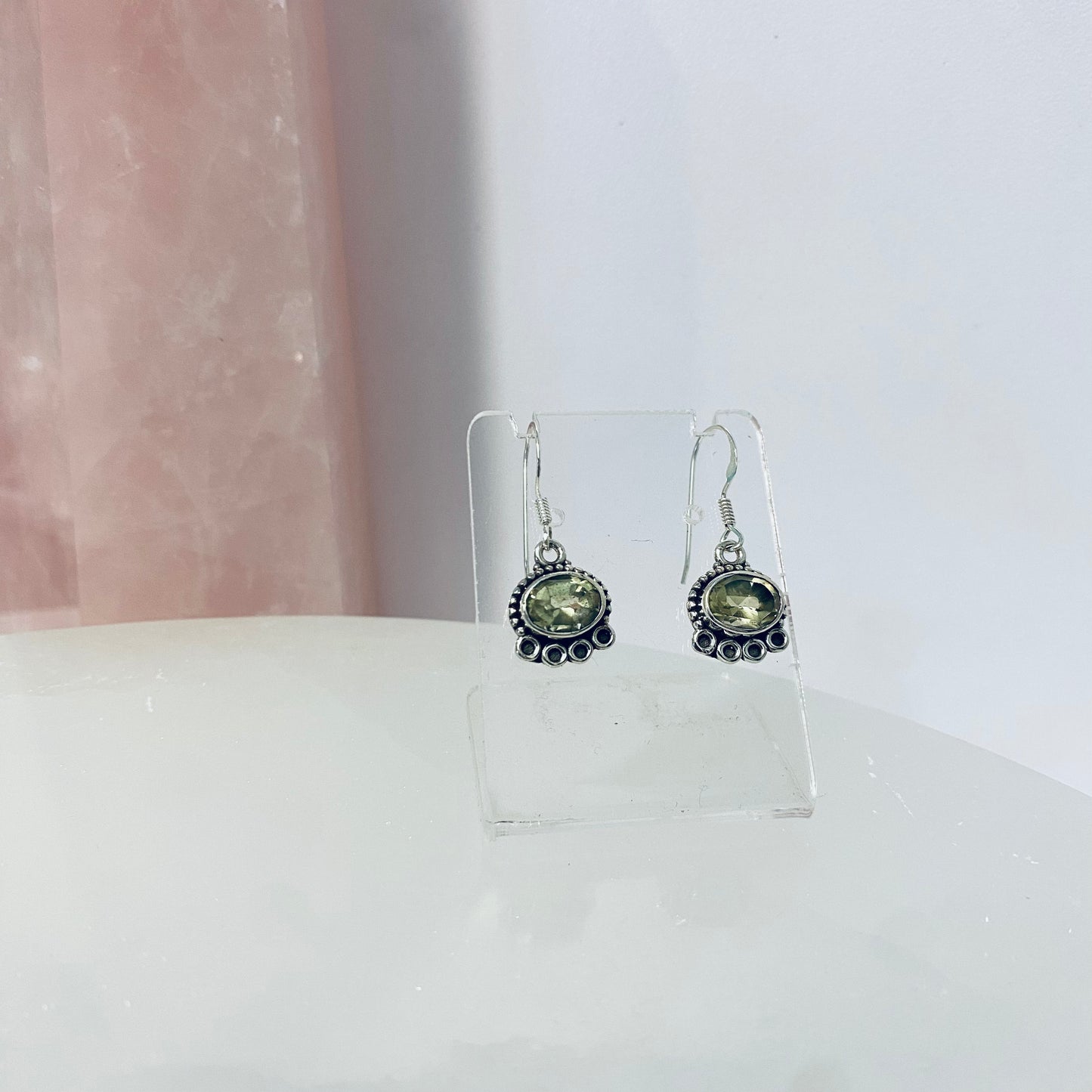 A pair of silver oval earrings with lemon quartz stones displayed on a stand against a pink background.