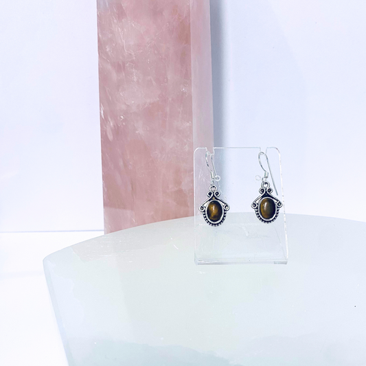 A pair of oval tiger eye earrings with silver accents displayed on a clear stand against a pink marble background.