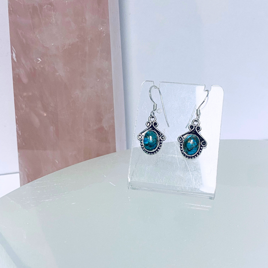 A pair of silver earrings featuring turquoise stones, displayed on a clear stand against a pink background.
