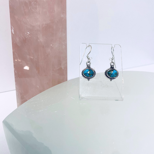 A pair of silver earrings with oval turquoise stones displayed on a clear stand against a pink crystal backdrop.