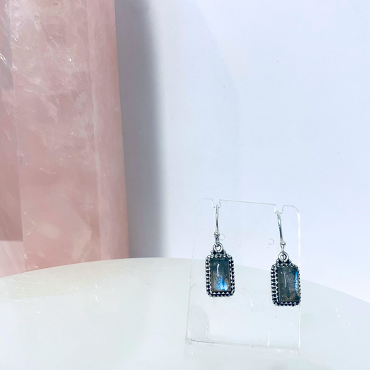 A pair of silver labradorite earrings displayed on a stand with a pink crystal in the background.