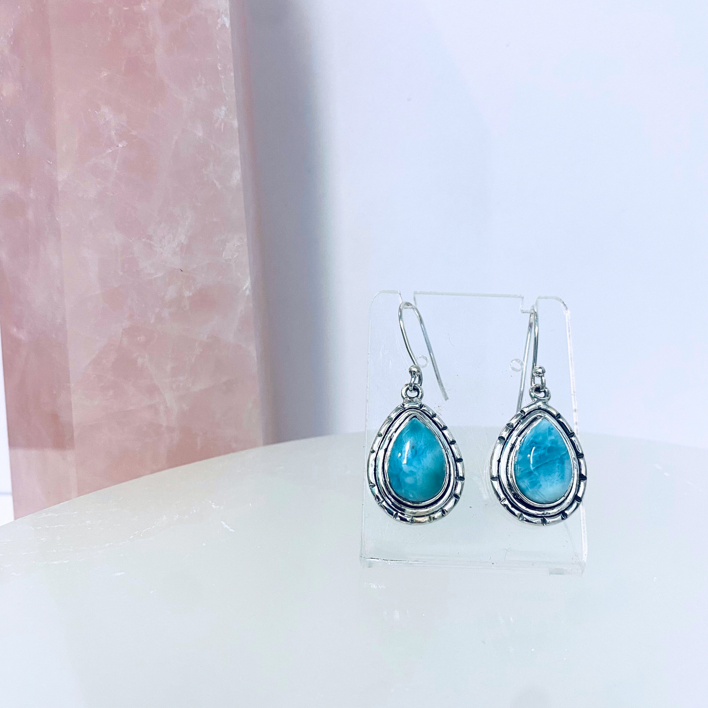 A pair of silver teardrop-shaped larimar earrings displayed on a clear stand against a pink-marbled background.