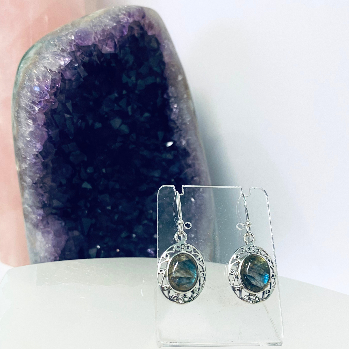 A pair of silver earrings featuring labradorite stones, displayed in front of an amethyst crystal.