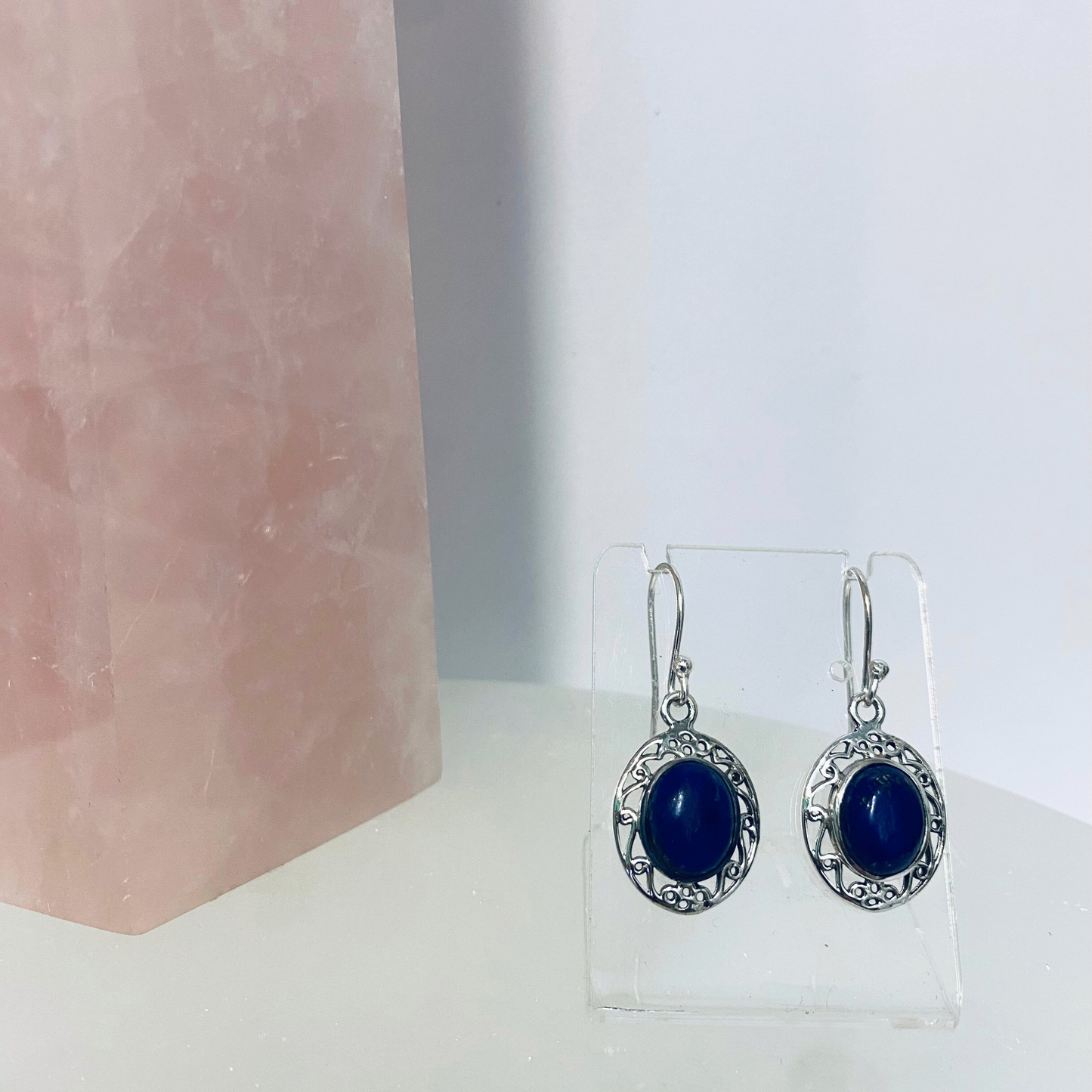 A pair of silver earrings featuring lapis lazuli stones, displayed against a white background with a pink crystal to the side.